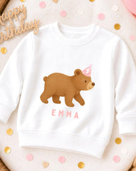 A white sweatshirt with a cartoon bear wearing a birthday hat and the name "Emma" printed on it, surrounded by confetti.