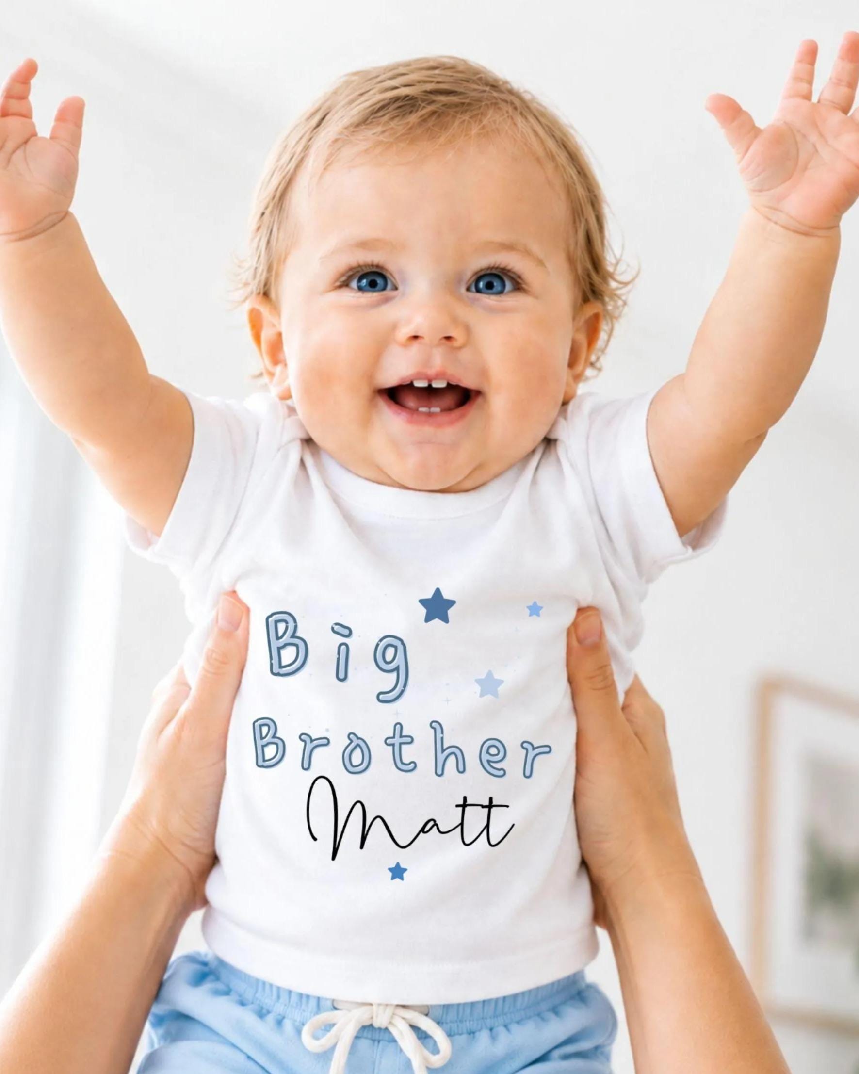 A smiling baby wearing a white shirt with the text "Big Brother Matt" and blue shorts, being held up by someone's arms.