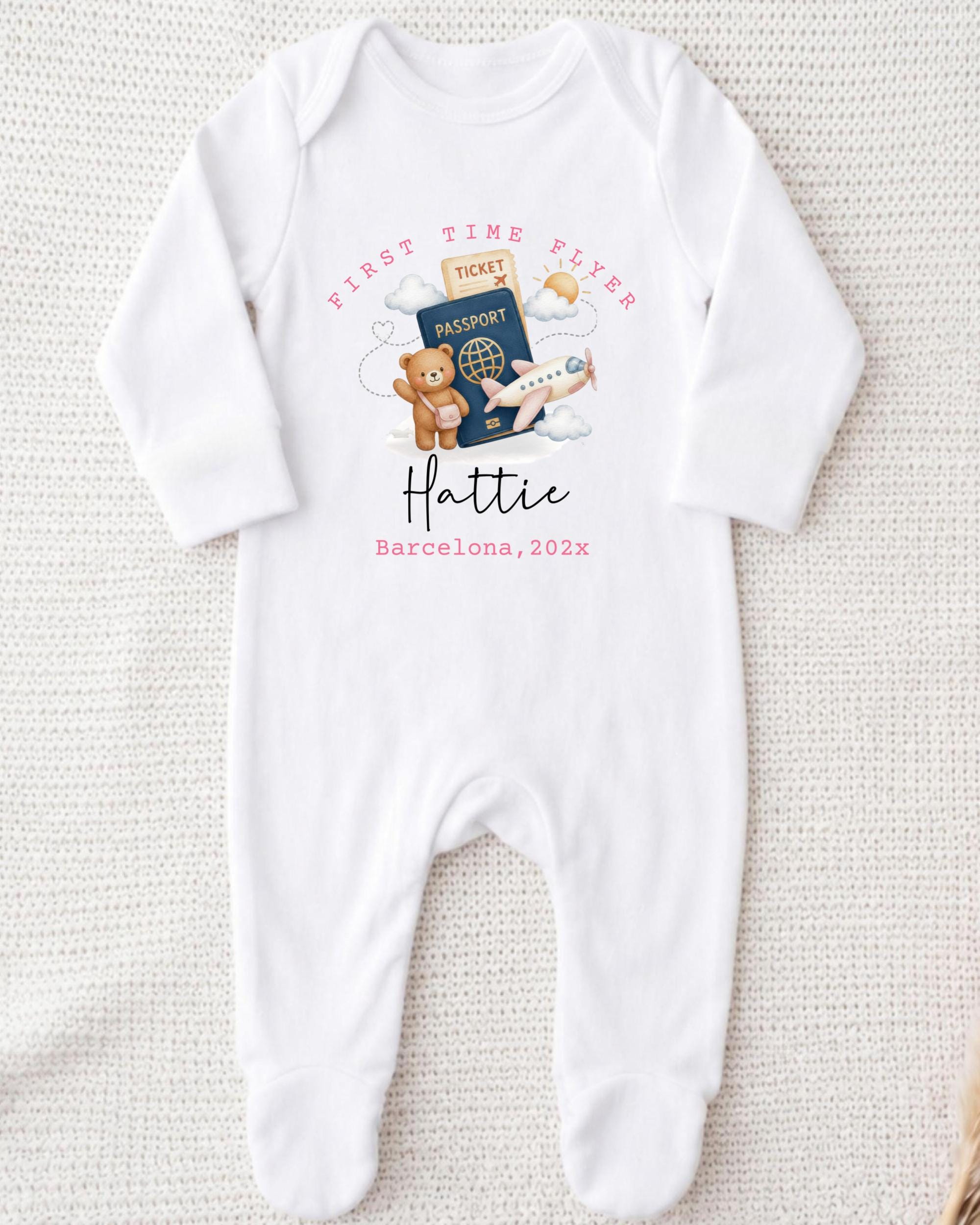 A white onesie with a graphic design on it, featuring a teddy bear, a passport, and an airplane. The text on the onesie says "First Time Flyer Hattie Barcelona, 202x".