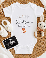 A baby announcement t-shirt with the text "BABY Wilson Coming Soon" and an image of a fox, along with a pair of white crocheted bunny slippers and a black and white baby announcement.