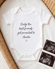 A white baby onesie with the text "Only the best friends get promoted to Auntie" printed on it, along with a heart symbol. The onesie is placed on a bed, and there are two black and white images of an ultrasound of a baby's belly visible in the background.