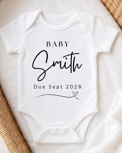 Personalisable white Baby Vest for a pregnancy announcement, printed in black with 'Baby Smith Due Sept 2028' and a fine-line heart.