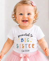 A smiling baby girl wearing a white t-shirt with text and a pink bow.
