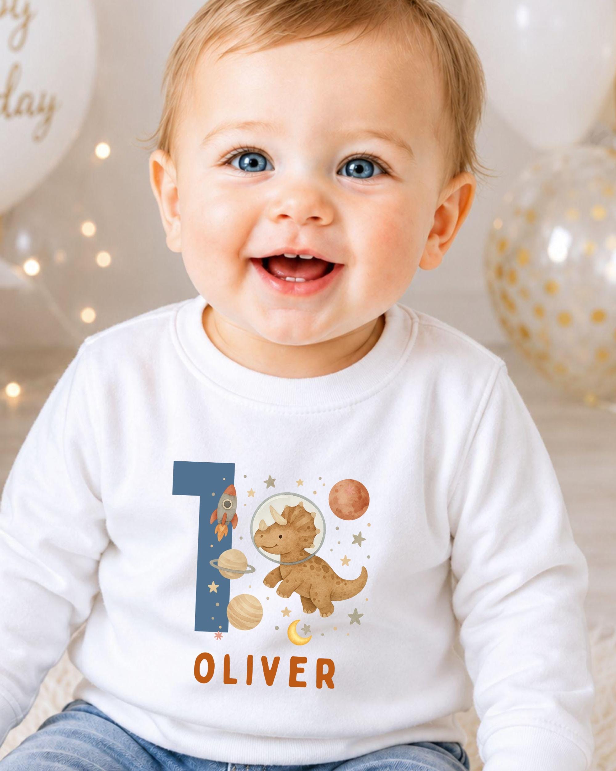 A smiling baby boy wearing a white shirt with a dinosaur and space-themed design, sitting in front of balloons.