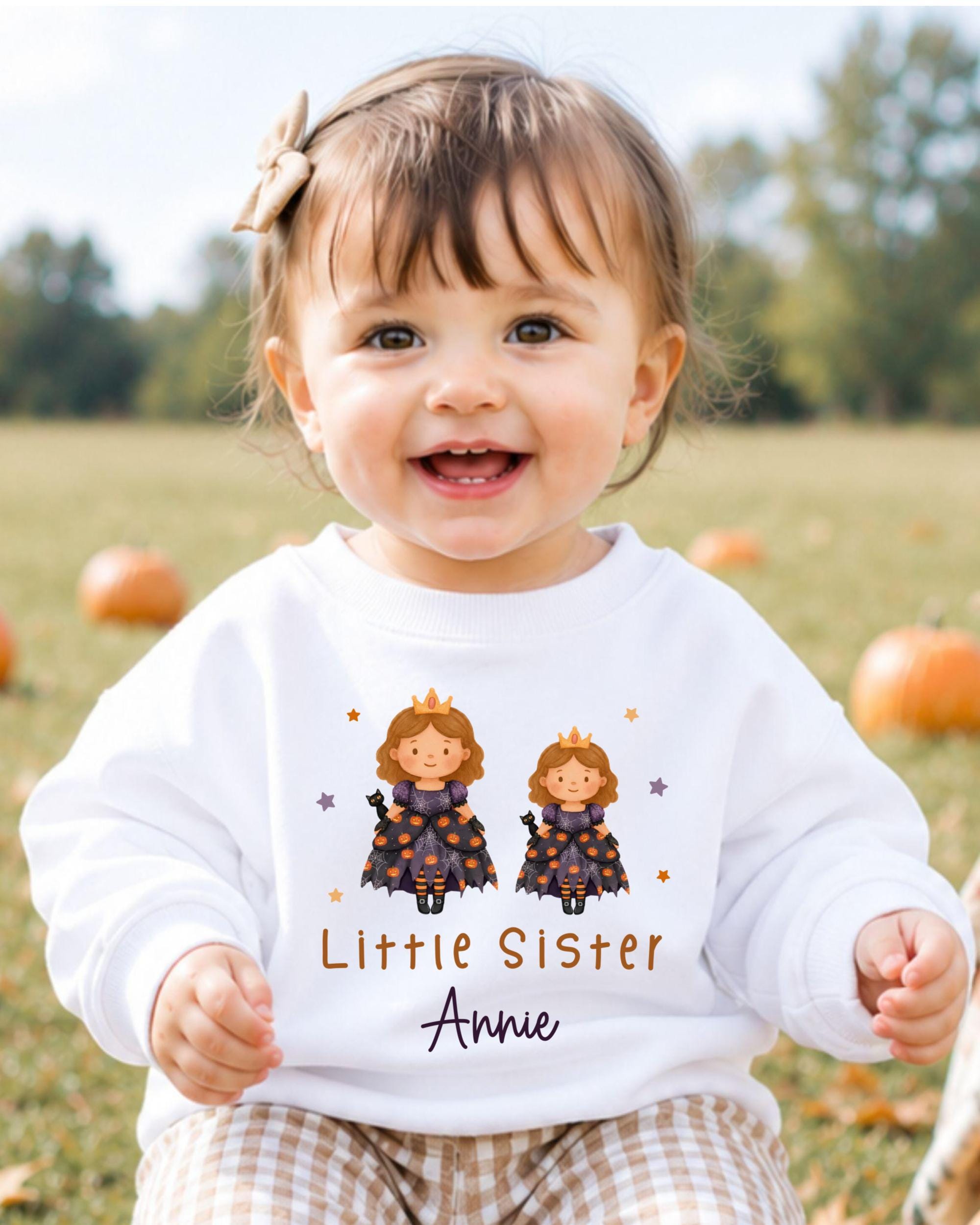 A smiling baby girl wearing a white sweater with a cartoon image of two princesses and the text "Little Sister Annie" printed on it.