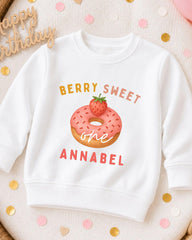 A white sweatshirt with a graphic of a strawberry donut and the text "Berry Sweet One Annabel" printed on it.
