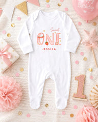 A white onesie with the text "Sweet One" and the name " Jessica" printed on it, along with a pink star and a pink flower, surrounded by confetti and other baby-themed decorations.