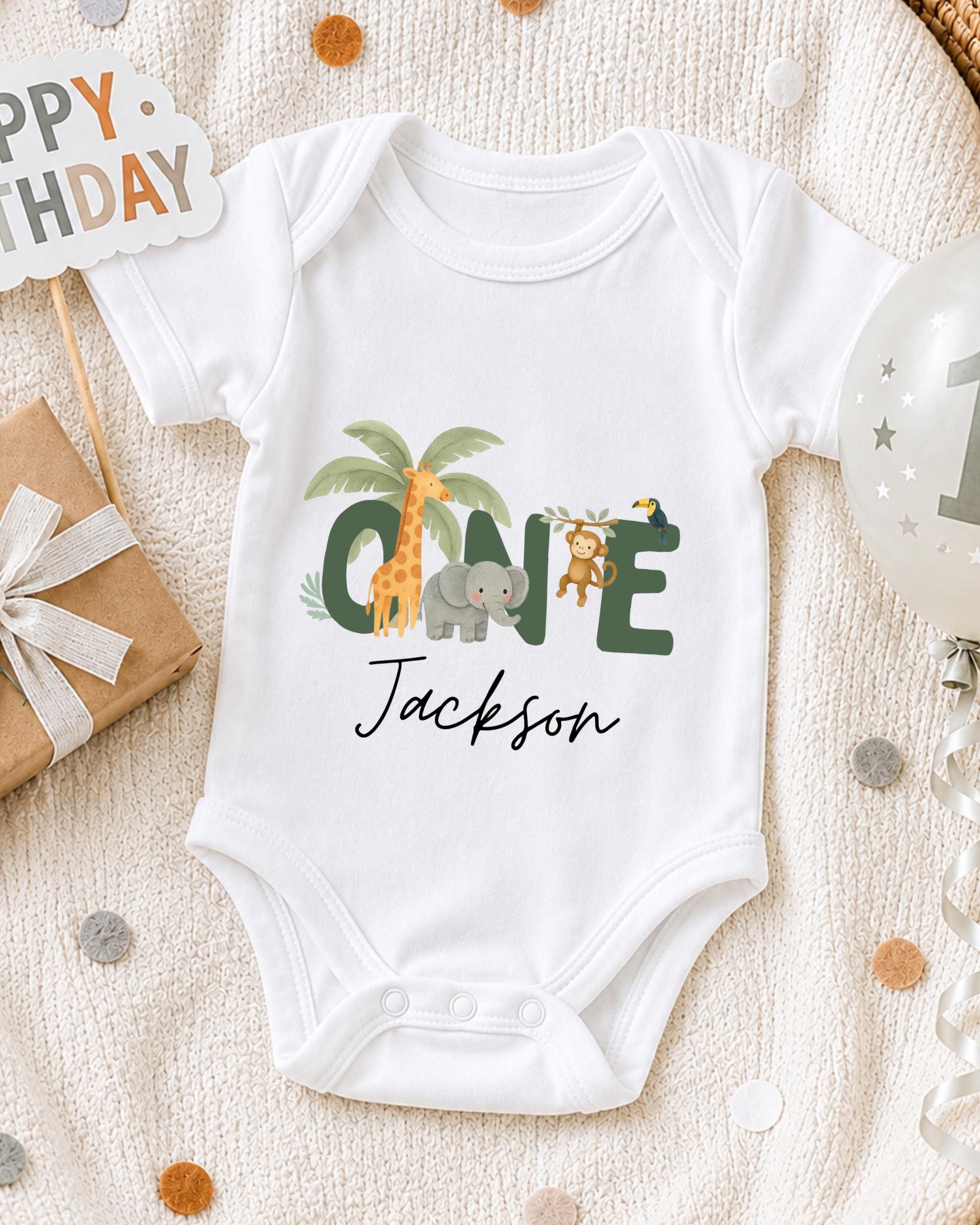 A white onesie with a personalized name and animal designs, including a giraffe, elephant, and monkey, is displayed on a blanket surrounded by birthday decorations and a "Happy Birthday" sign.