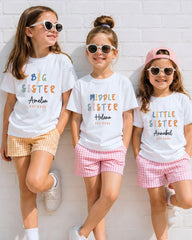 Three young girls wearing white t-shirts with text and sunglasses are standing against a white brick wall.