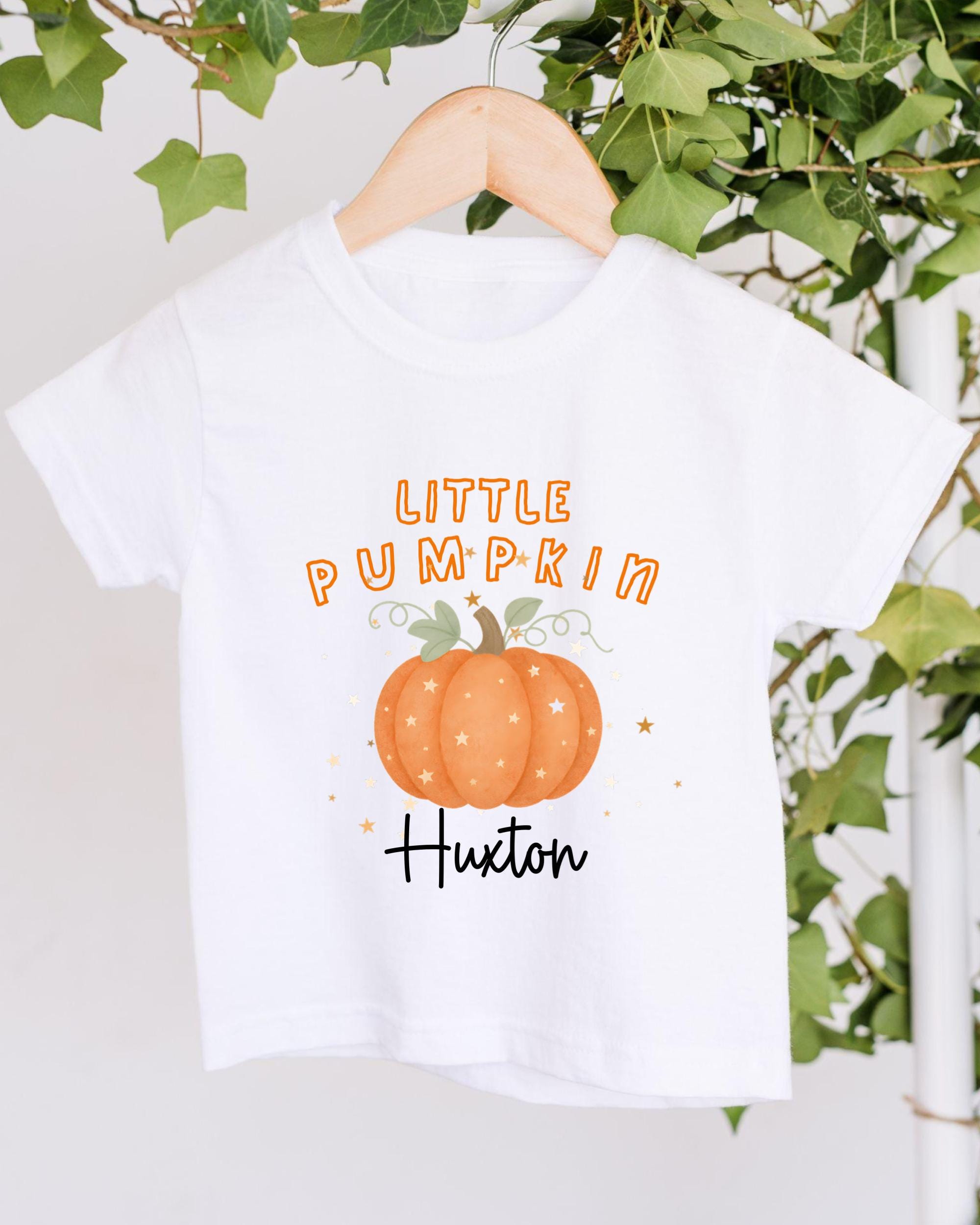 A white t-shirt with a pumpkin graphic and text printed on it, hanging on a wooden hanger.