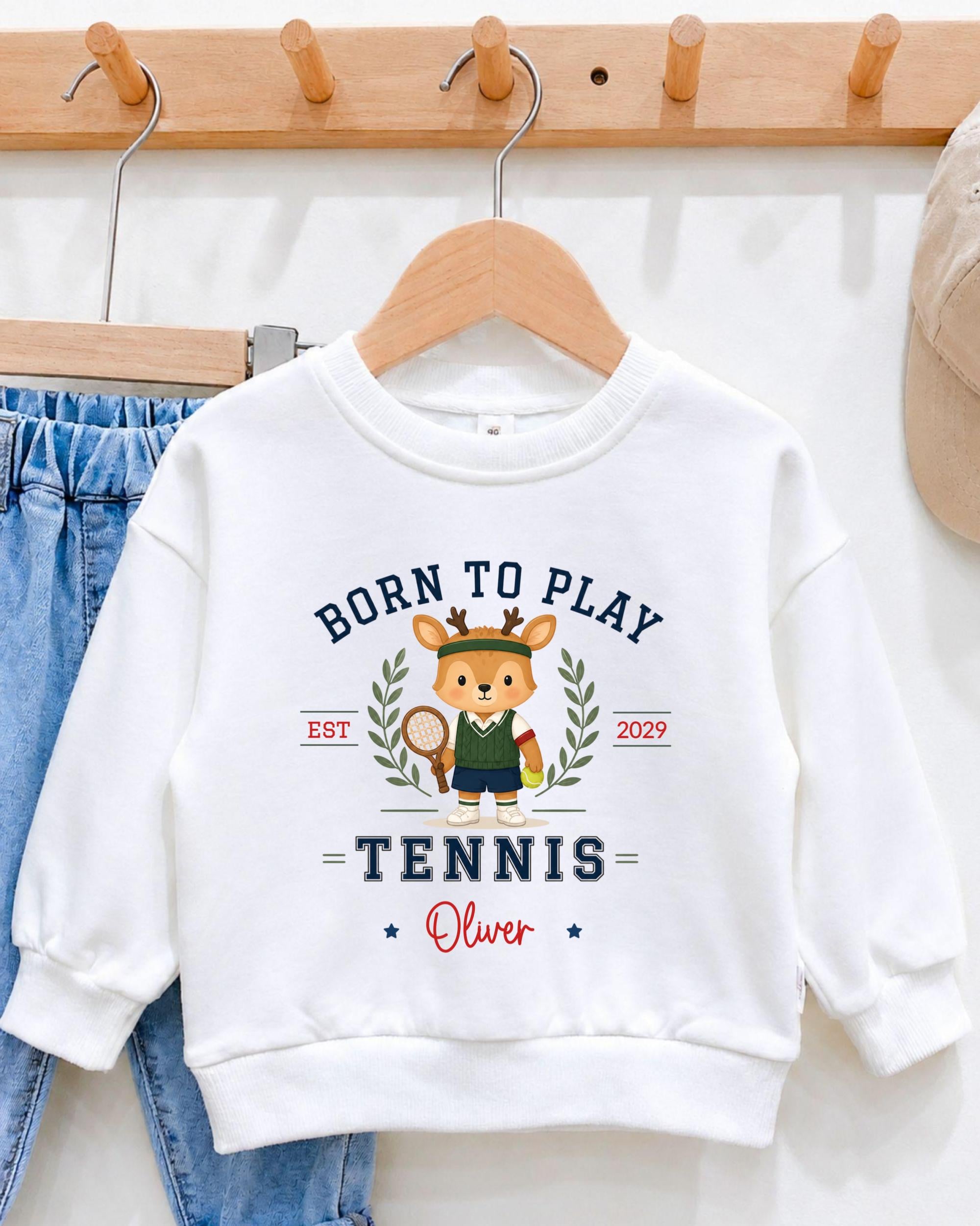 A white sweatshirt with a cartoon deer wearing a tennis outfit and the text "Born to play tennis Oliver" printed on it.