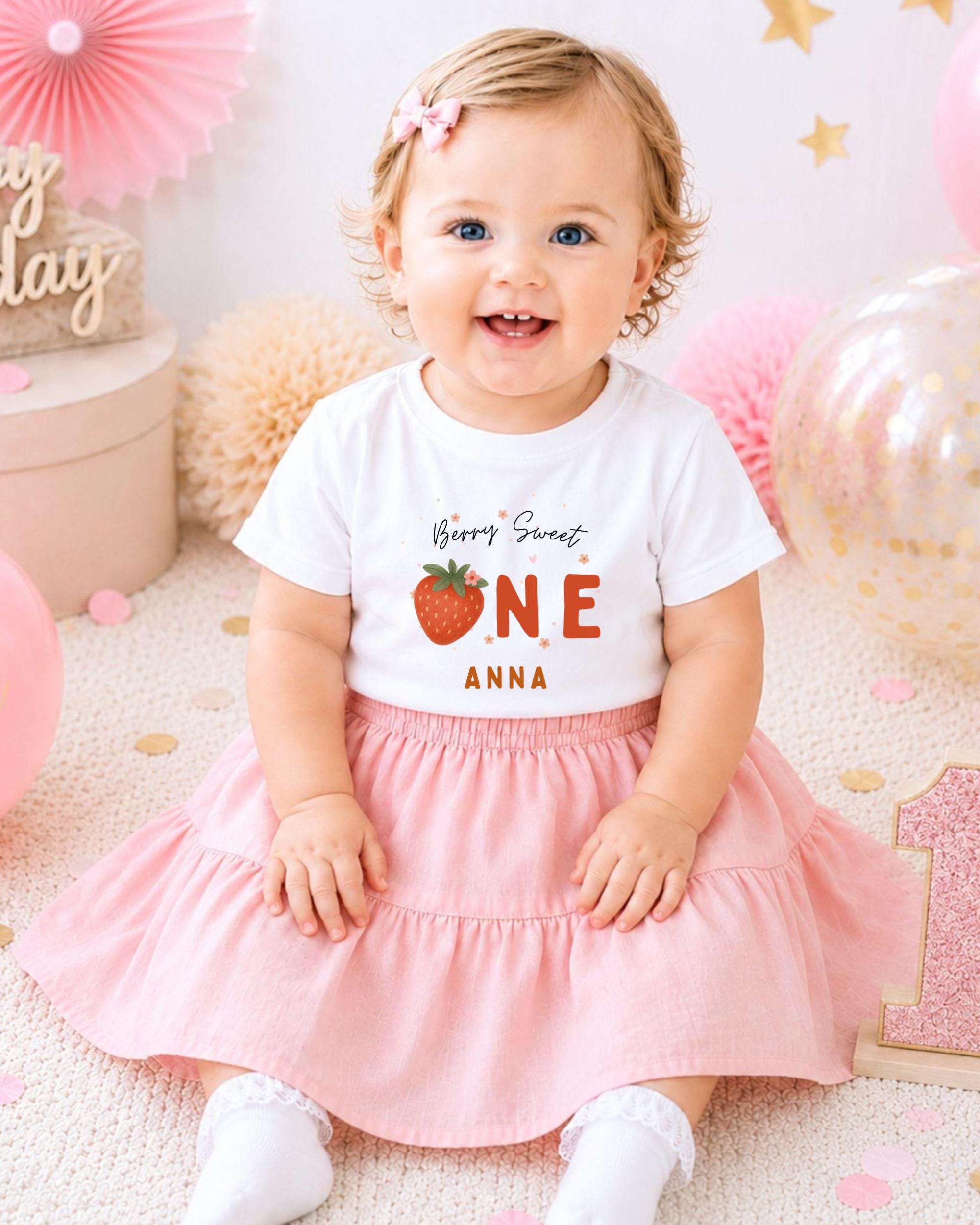 A smiling baby girl wearing a white t-shirt with a strawberry graphic and the text "Berry Sweet One Anna" sits on a pink and white background with balloons and confetti.