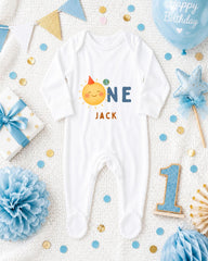 A white onesie with a sun graphic and the name "Jack" printed on it, surrounded by birthday decorations such as a blue balloon, blue and gold confetti, and a blue and white polka dot gift box.