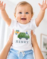 A smiling baby wearing a white shirt with a green truck and balloons on it, being held up by someone's arms.
