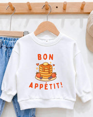 A white sweatshirt with a graphic of pancakes and the text " bon appétit!" hanging on a wooden hanger.