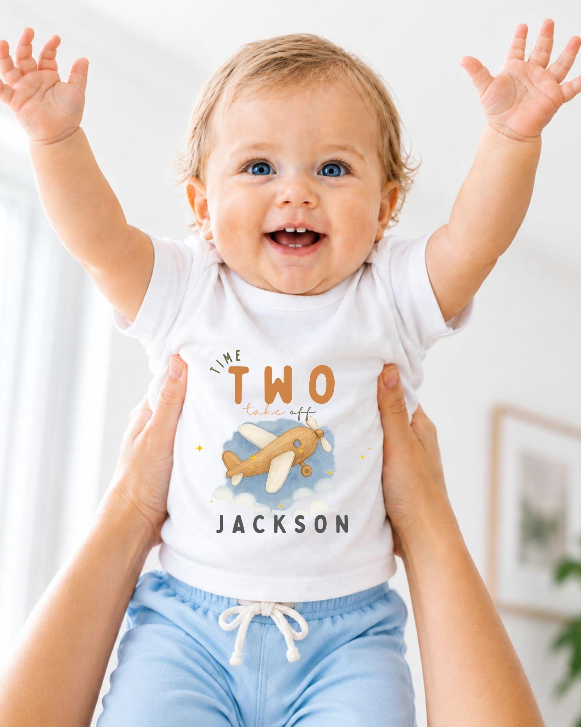 A smiling baby wearing a white t-shirt with an airplane graphic and the text "The two take off Jackson" is being held up by someone's arms.