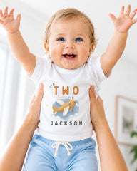 A smiling baby wearing a white t-shirt with an airplane graphic and the text "The two take off Jackson" is being held up by someone's arms.