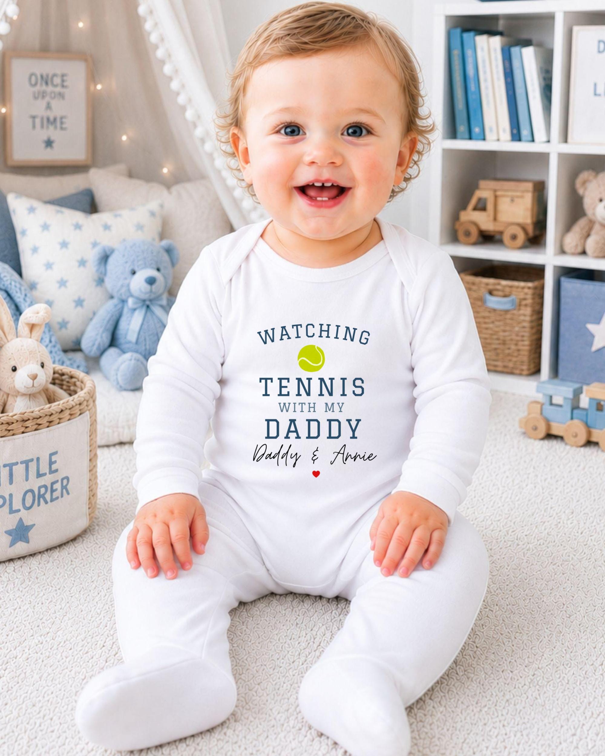 A smiling baby boy wearing a white onesie with the text "Watching Tennis with my Daddy Daddy & Annie" is sitting on the floor in a nursery.