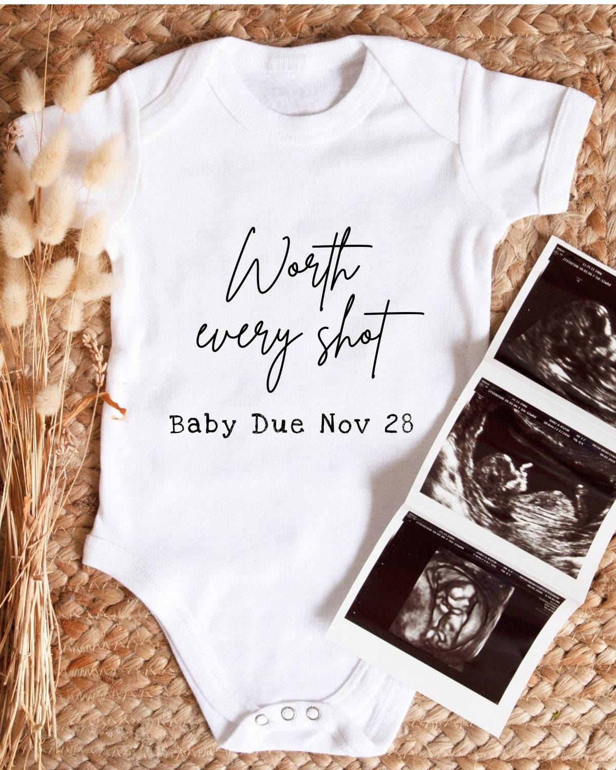 A white baby onesie with the text " Worth every shot Baby Due Nov 28" printed on it, along with an image of an ultrasound.