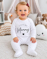 A smiling baby girl wearing a white onesie with the text "I love my Mummy" sits on a white carpet surrounded by stuffed animals.