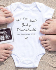 a white onesie with text and an image of an Ultrasound scan of a baby, surrounded by hands on a sandy beach.