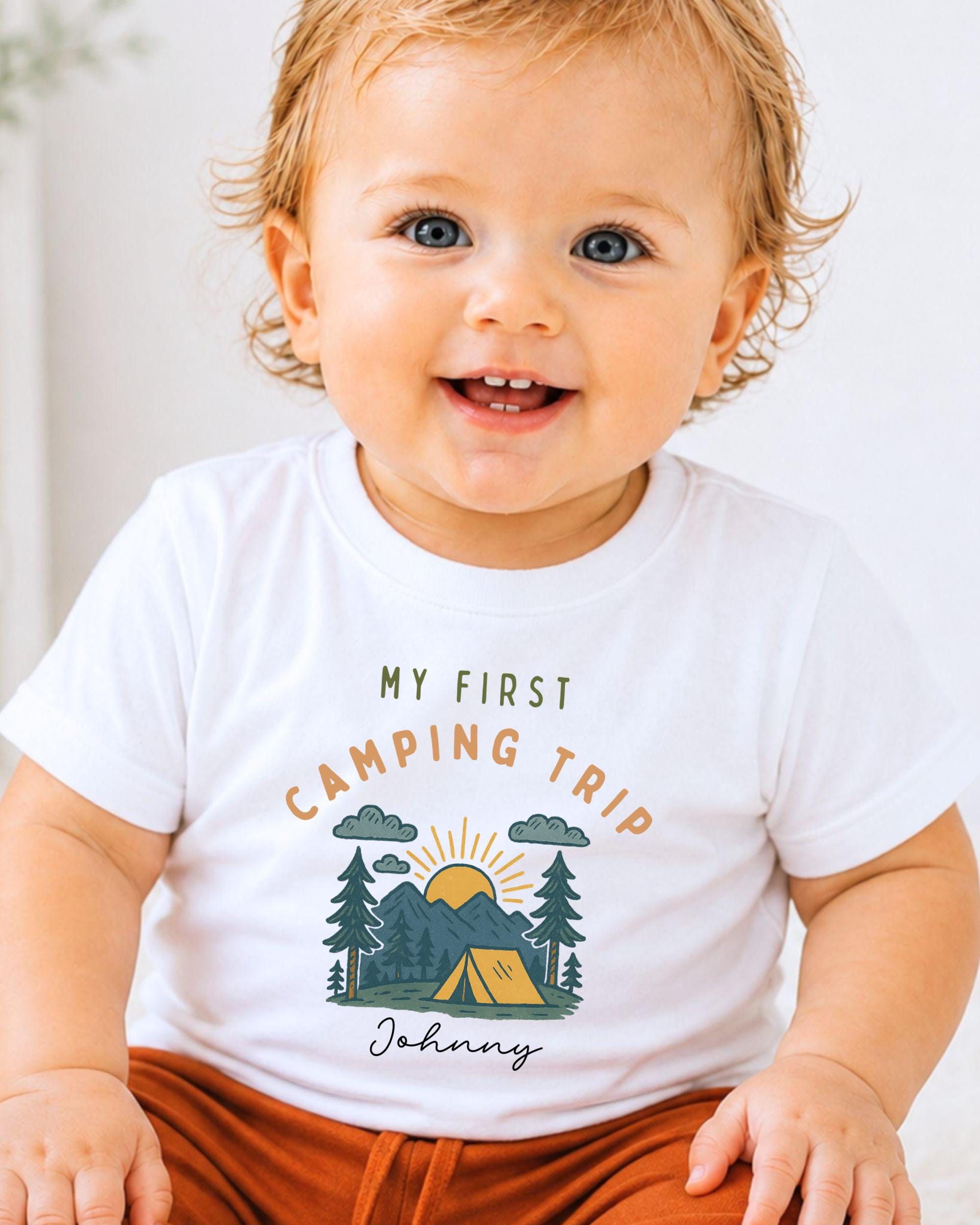 A smiling baby wearing a white t-shirt with a camping-themed graphic and text.