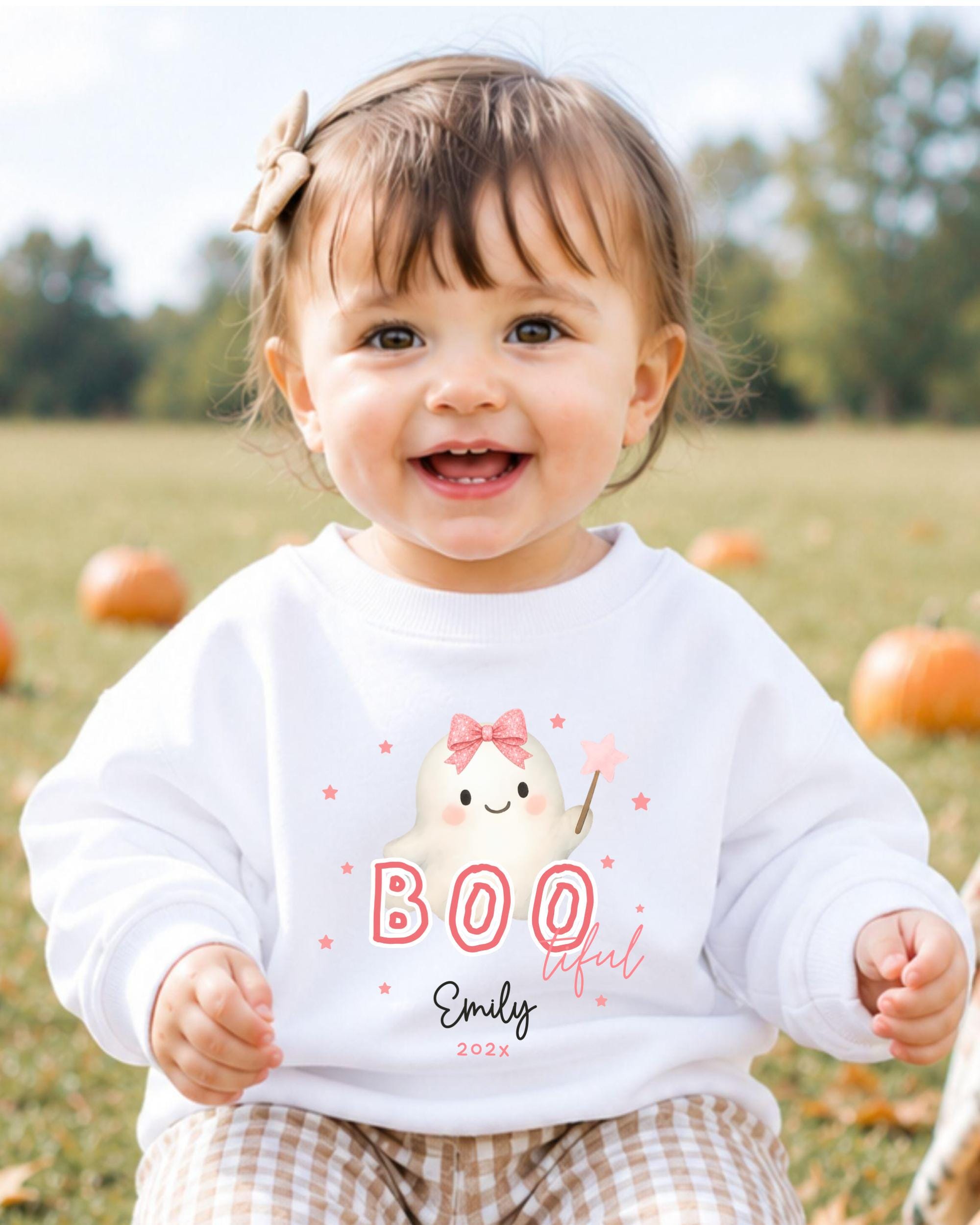 A smiling baby girl wearing a white sweater with a ghost and the text "BOO lifer" printed on it, sitting in a field with pumpkins in the background.