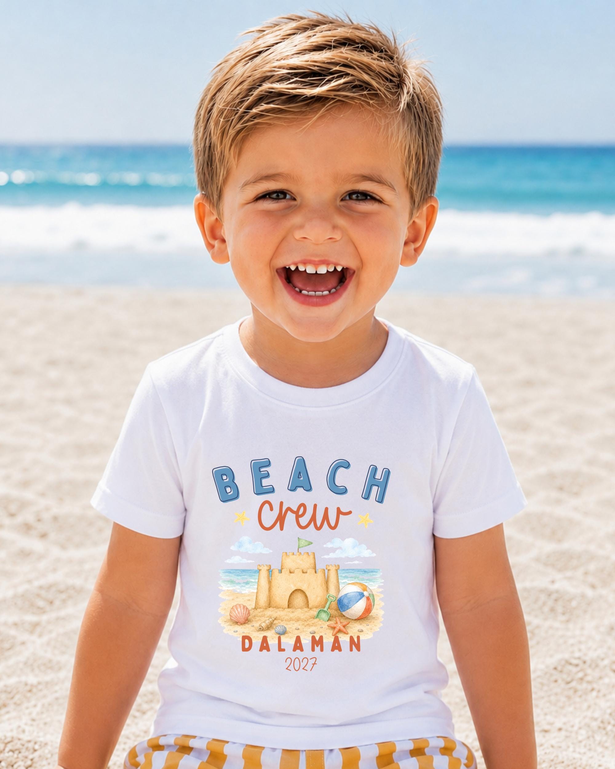 A young boy with a big smile on his face, wearing a white t-shirt with a beach-themed design, is standing on a sandy beach with the ocean in the background.