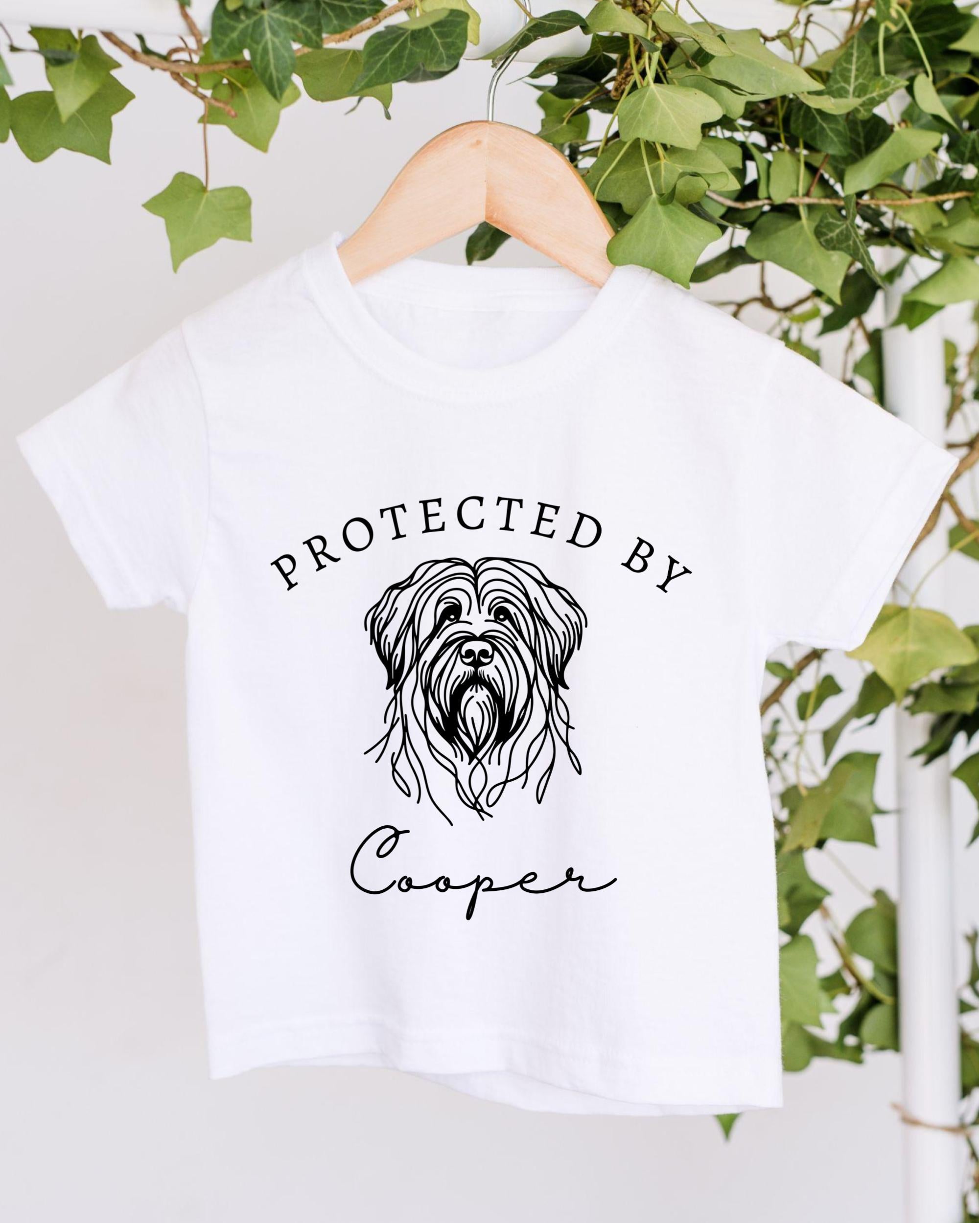 A white t-shirt with a graphic of a dog's head and the text "PROTECTED BY Cooper" printed on it, hanging on a wooden hanger.