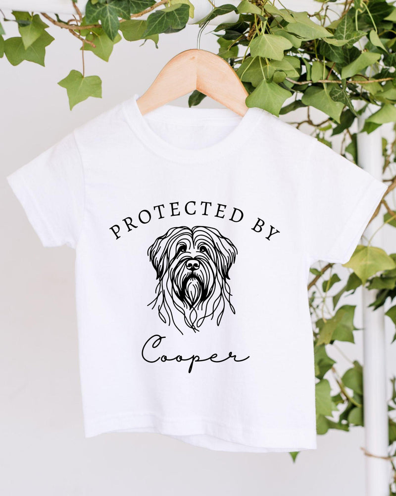 A white t-shirt with a graphic of a dog's head and the text "PROTECTED BY Cooper" printed on it, hanging on a wooden hanger.