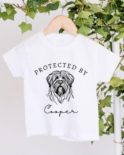 A white t-shirt with a graphic of a dog's head and the text "PROTECTED BY Cooper" printed on it, hanging on a wooden hanger.