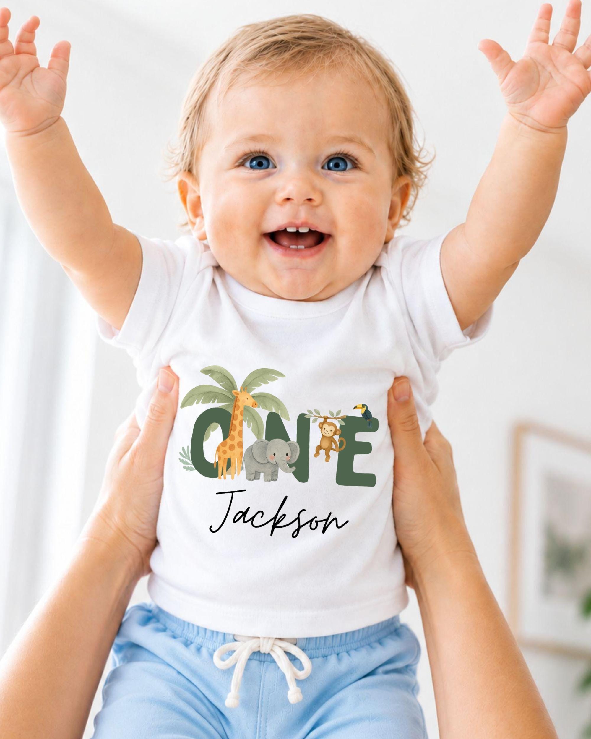 A smiling baby wearing a white t-shirt with a personalized name and animal print, being held up by someone's arms.