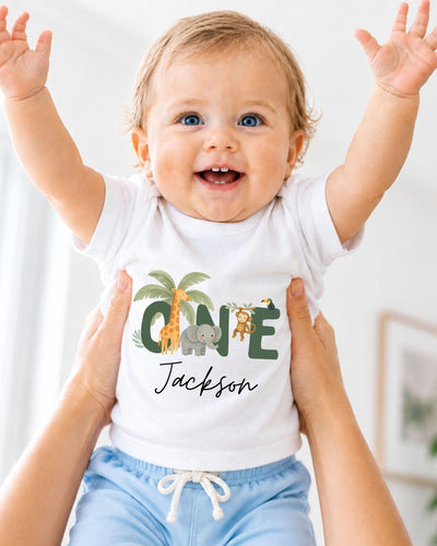 A smiling baby wearing a white t-shirt with a personalized name and animal print, being held up by someone's arms.