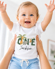 A smiling baby wearing a white t-shirt with a personalized name and animal print, being held up by someone's arms.