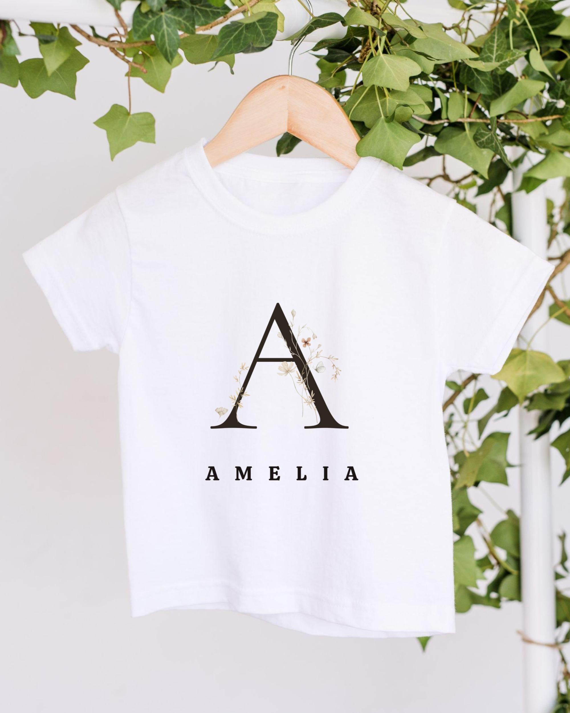 A white t-shirt with a monogrammed letter "A" and the name "Amelia" printed on it, hanging on a wooden hanger.
