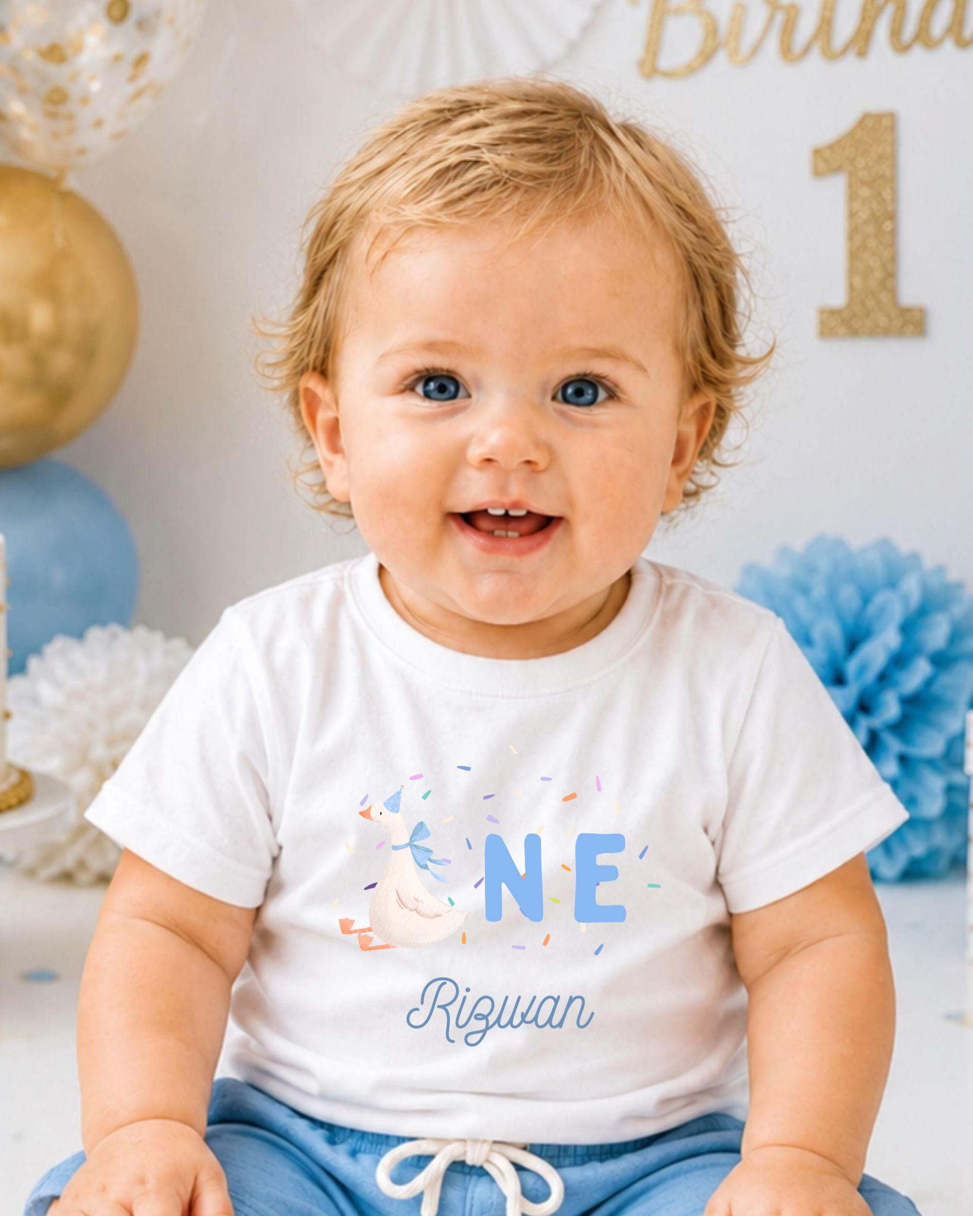 A smiling baby boy wearing a white t-shirt with a blue design and the word "Rigwan" printed on it, sitting in front of a birthday party backdrop.