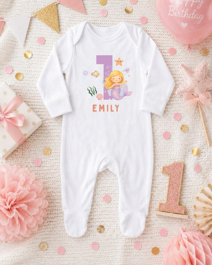 A white bodysuit with a mermaid design and the name "EMILY" printed on it, surrounded by pink and gold decorations, including a pink star, a pink flower, and confetti.