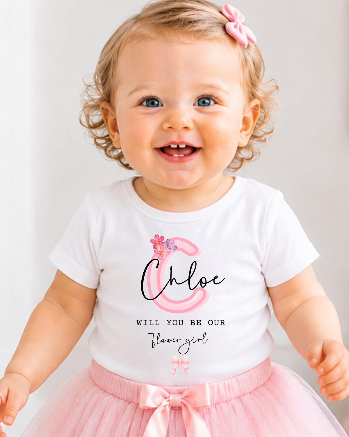 A smiling baby girl wearing a white t-shirt with a personalized monogram and a pink tutu skirt.