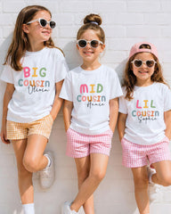 Three young girls wearing white t-shirts with colorful text and sunglasses, standing against a white brick wall.