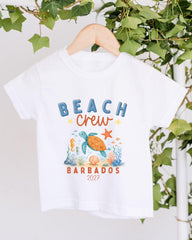 A white t-shirt with a graphic design featuring a turtle, starfish, and coral, along with the text "BEACH CREW" and "BARBADOS 2027".