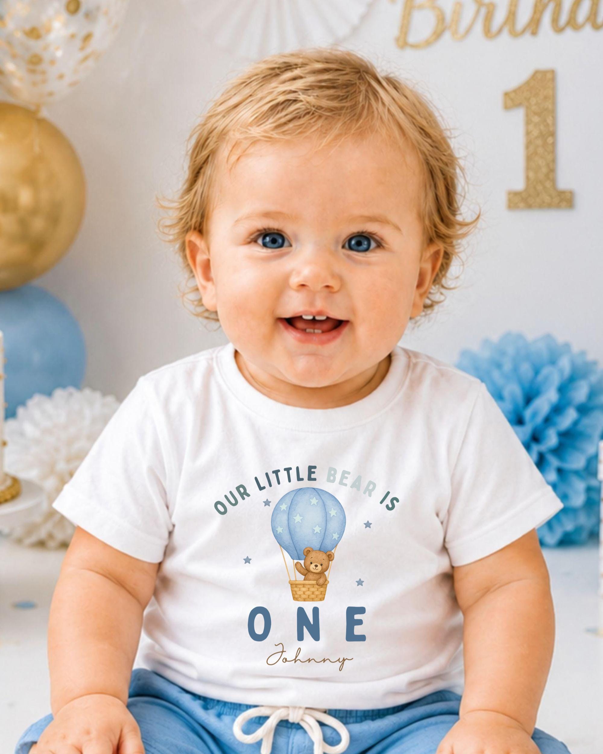 A smiling baby boy wearing a white t-shirt with a cartoon image of a bear and the text "Our little bear is 1" is sitting in front of a birthday party decoration.