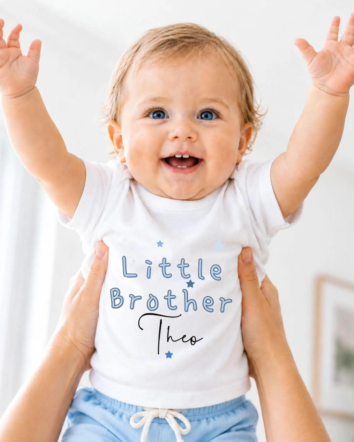 A smiling baby wearing a white shirt with the text "Little Brother Tho" is being held up by someone's arms.