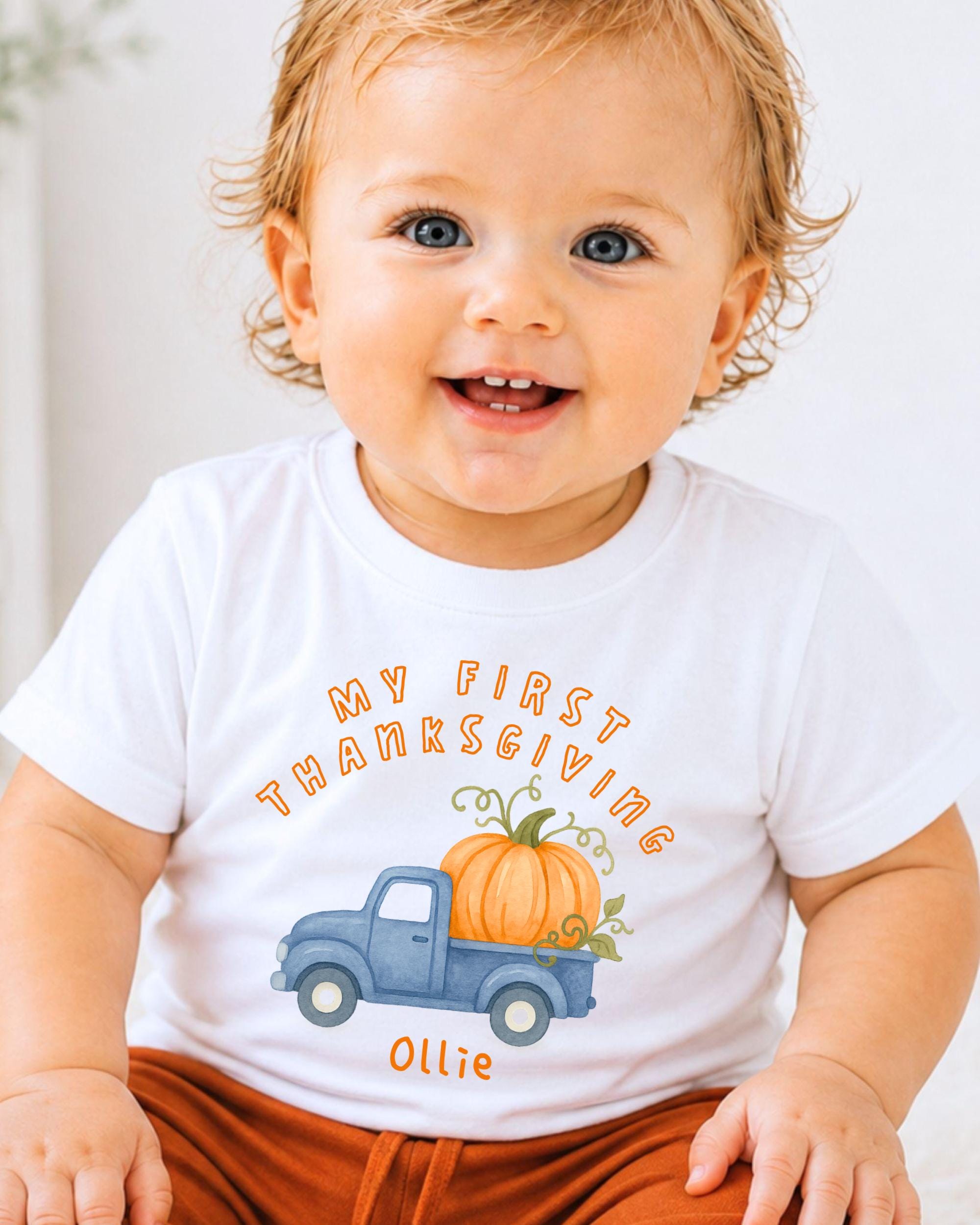A smiling baby boy wearing a white t-shirt with a graphic of a blue truck carrying a pumpkin and the text "My first Thanksgiving Ollie".