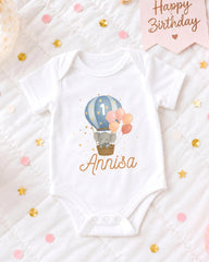A white onesie with a cartoon elephant and balloons, along with the name "Anisa" printed on it, is displayed on a bed with confetti and a birthday card.