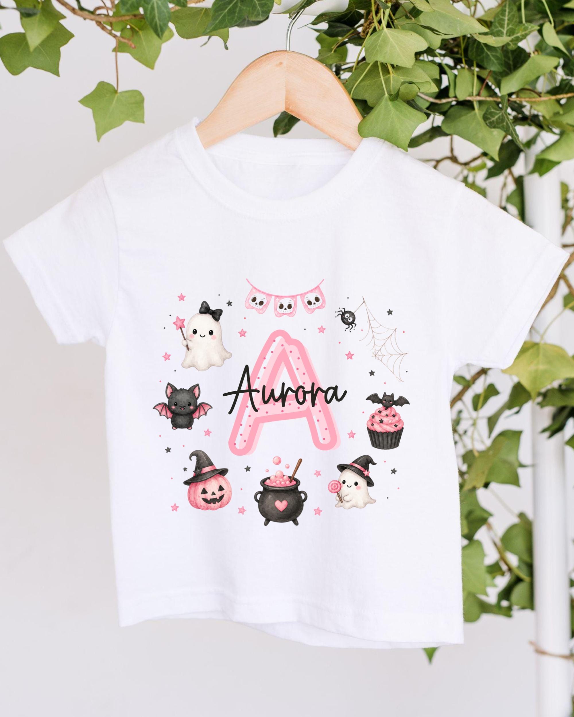 A white t-shirt with a personalized design featuring a pink "A" surrounded by various Halloween-themed illustrations, including a ghost, bats, a spider, a pumpkin, a witch's hat, a pumpkin, a cupcake, and a spider web.