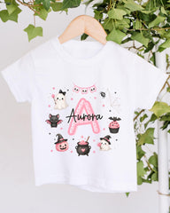 A white t-shirt with a personalized design featuring a pink "A" surrounded by various Halloween-themed illustrations, including a ghost, bats, a spider, a pumpkin, a witch's hat, a pumpkin, a cupcake, and a spider web.