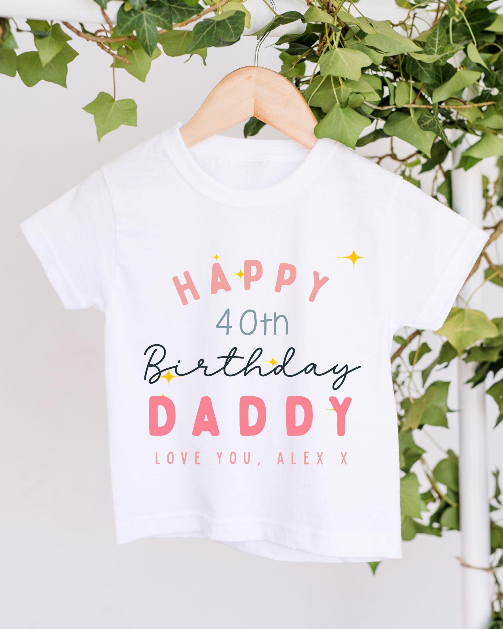 A white t-shirt with text and graphics hanging on a wooden hanger, with greenery in the background.