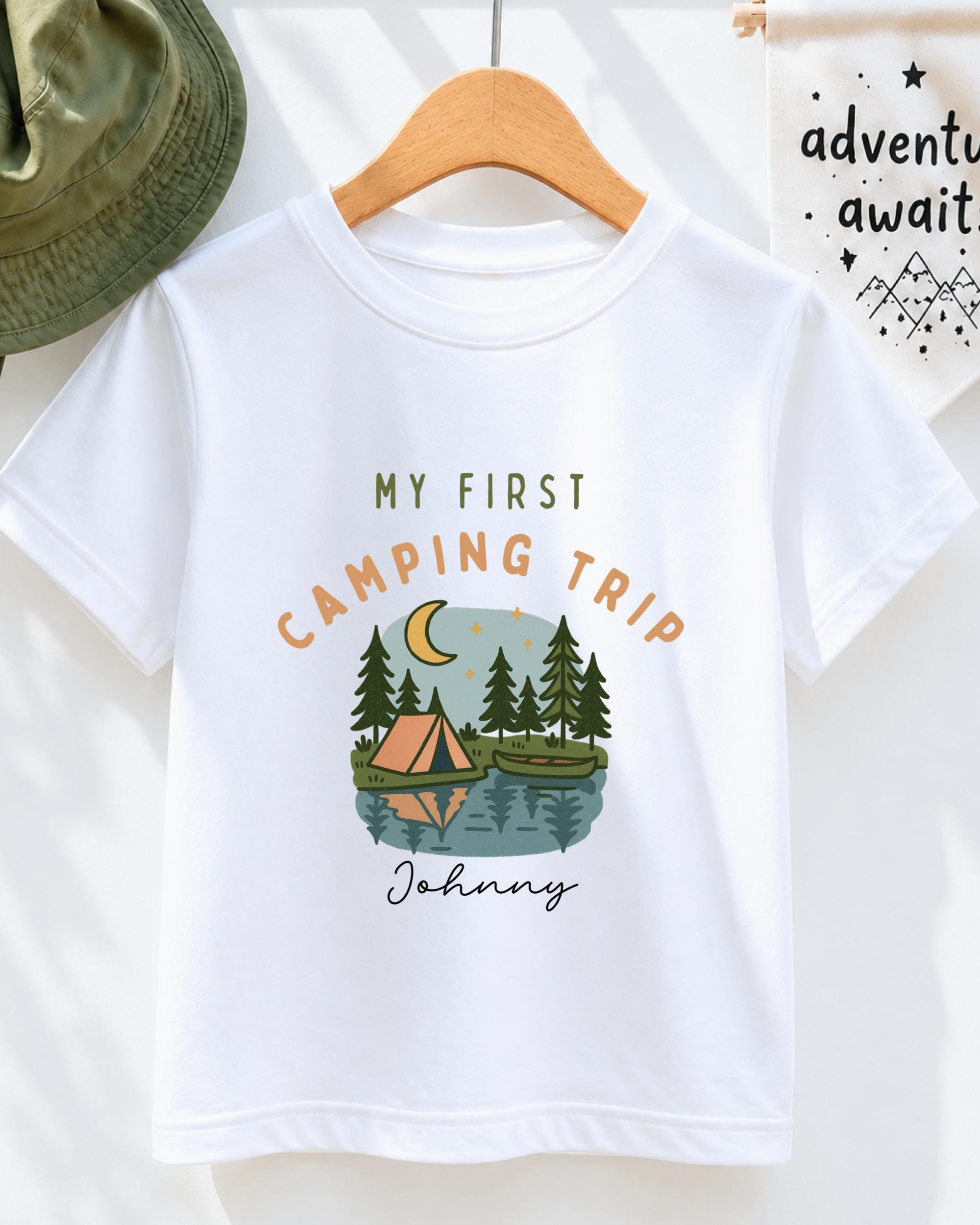 A white t-shirt with a graphic design depicting a camping scene with a tent, trees, and a lake, along with the text "My first camping trip" and "Johny" printed on it.