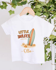 A white t-shirt with a graphic design featuring a surfboard, seashells, and a starfish, along with the text "Little Surfer - Ollie" printed on it.