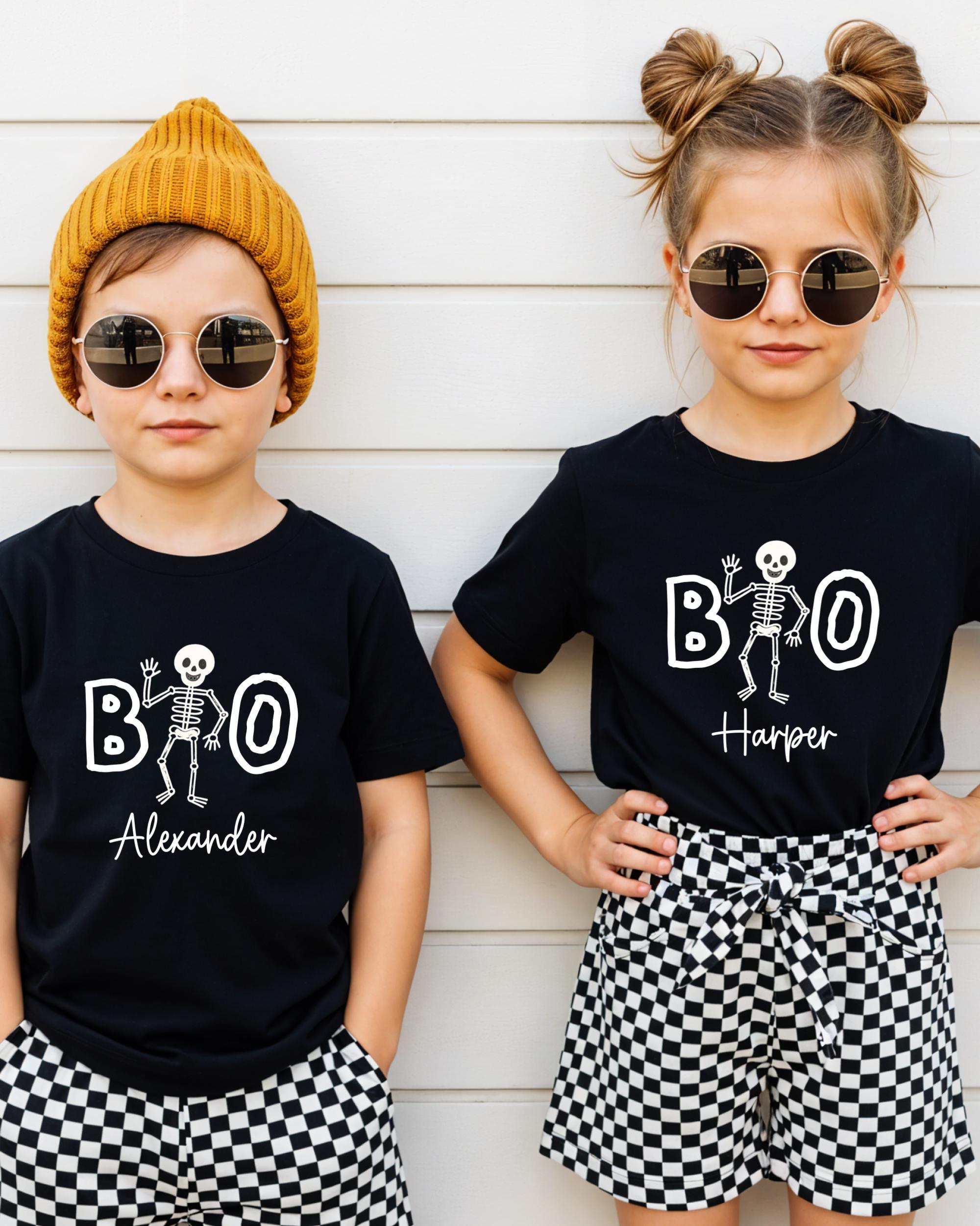 Two children wearing black t-shirts with a skeleton design and the text "BOO Harper" printed on them, along with round sunglasses and a yellow beanie.